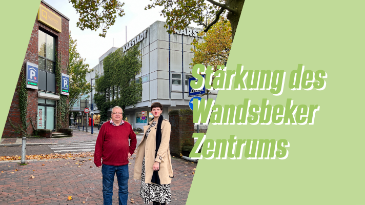 Foto von Ingo (SPD) und Julia B. (GRÜNE) vor dem Wandsbeker Quarree und Karstadt Gebäude. Ingo trägt einen roten Pulli, Julia einen beigen Trechcoat, beide stehen auf geplasterter Fläche, zwischen den Gebäuden verläuft eine kleine Straße.